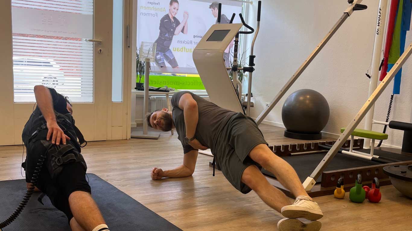 Zwei Männer beim Plank-Training in einem Fitnessstudio mit hellem Boden und Geräten.
